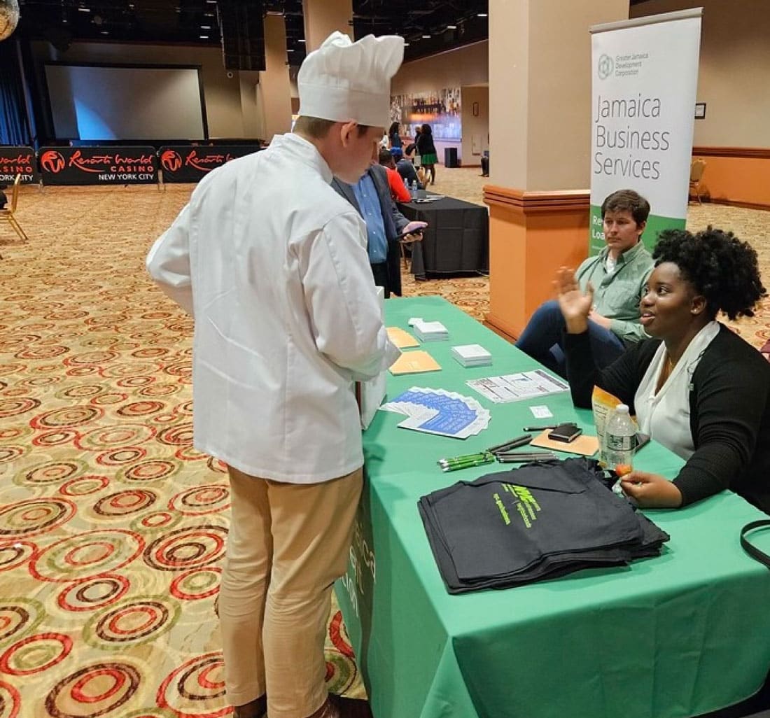 A career fair exhibit table with a banner displaying "Jamaica Business Services". A person in a chef's uniform is talking with a woman at the table.