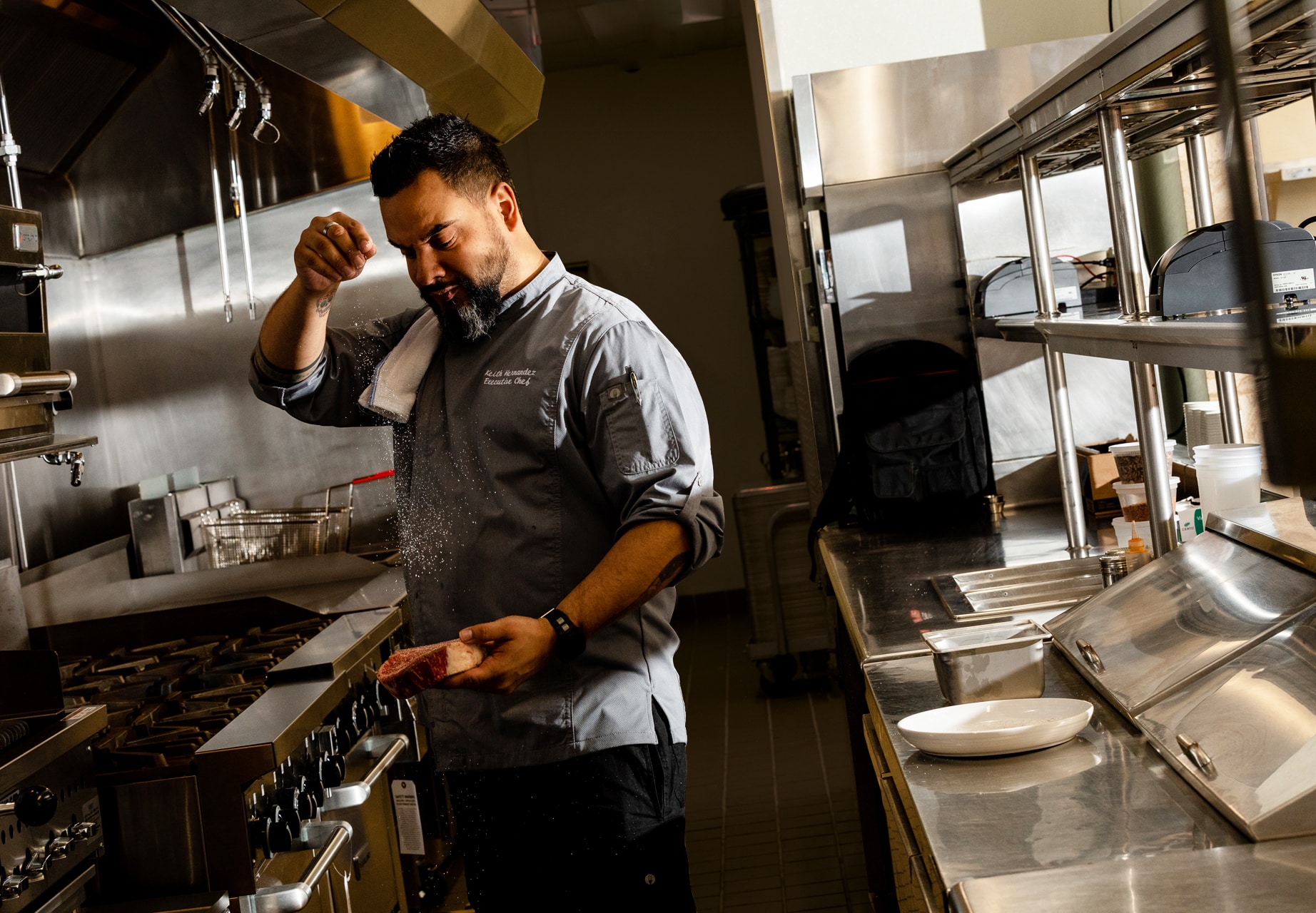A chef in a kitchen, wearing a white uniform, preparing food on a counter with stainless steel appliances behind him.