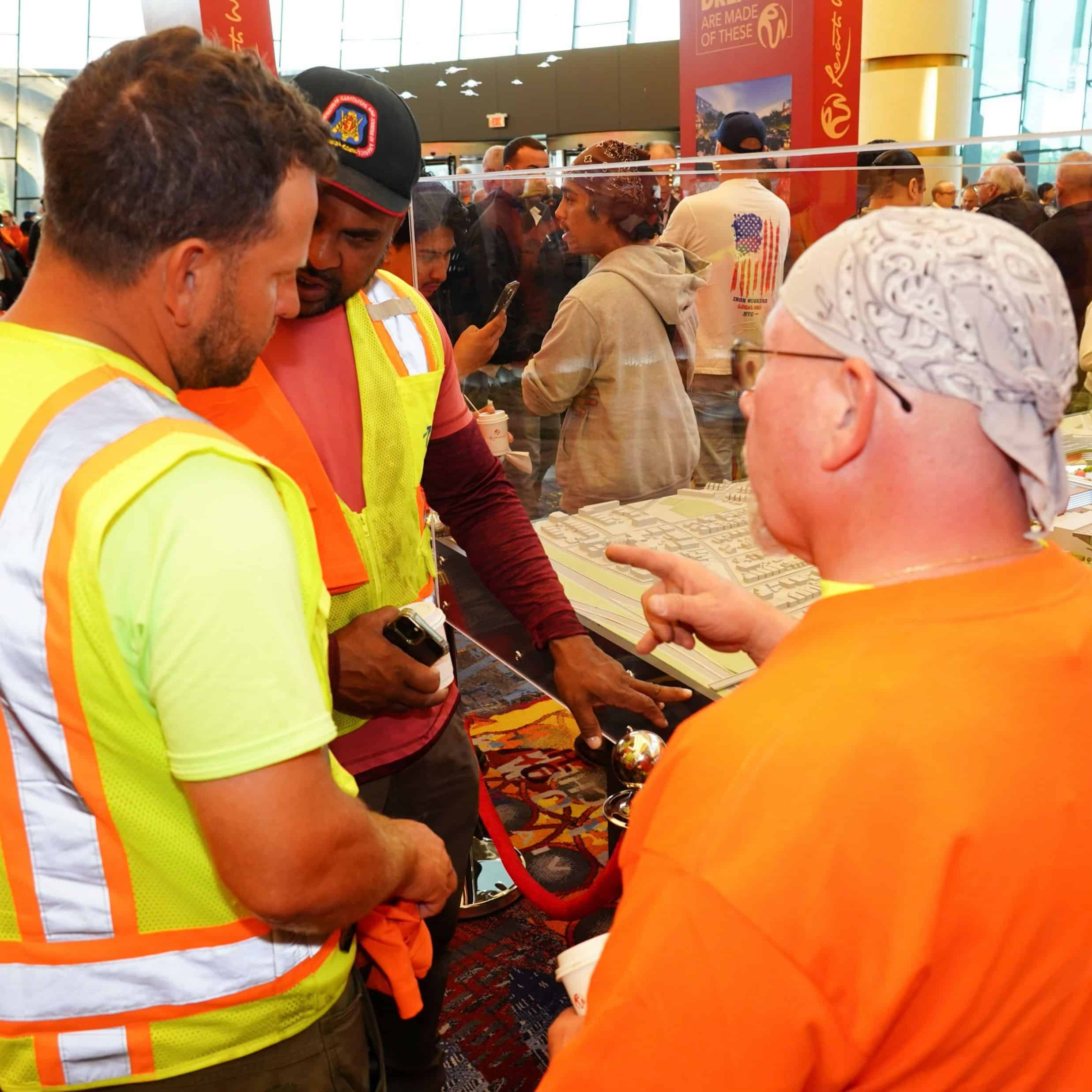 A construction worker in a safety vest and hardhat assisting travelers with luggage and documents at an airport security checkpoint.