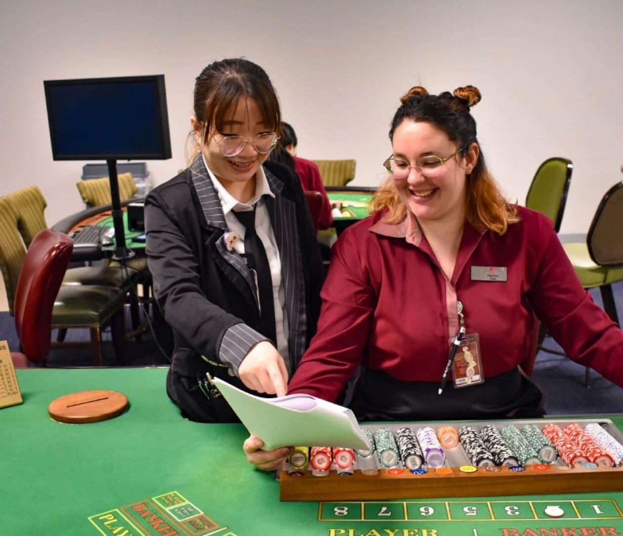 Two women at a casino gaming table reviewing a tablet device and discussing game materials.
