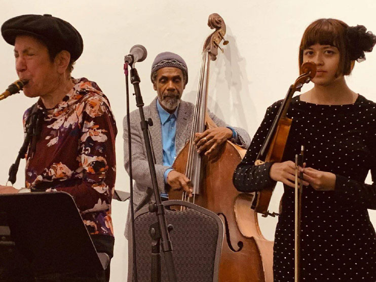 Three musicians on stage: a woman with an electric guitar, a man playing an upright bass, and a man singing into a microphone.