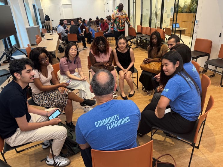 A diverse group of people sitting in a circle, with one person wearing a "Community Teamworks" shirt, engaging in a discussion.