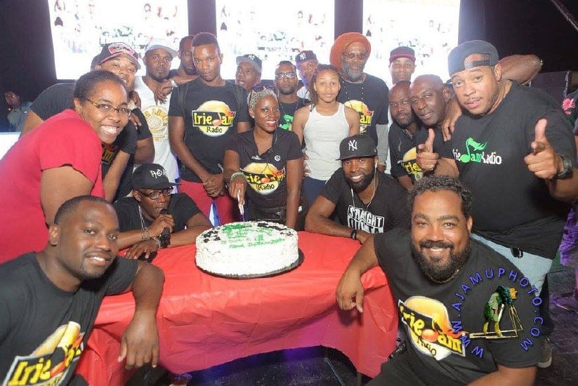 Group of people gathered around a birthday cake, wearing t-shirts that say "Irie Radio" and other slogans, smiling and giving thumbs up.