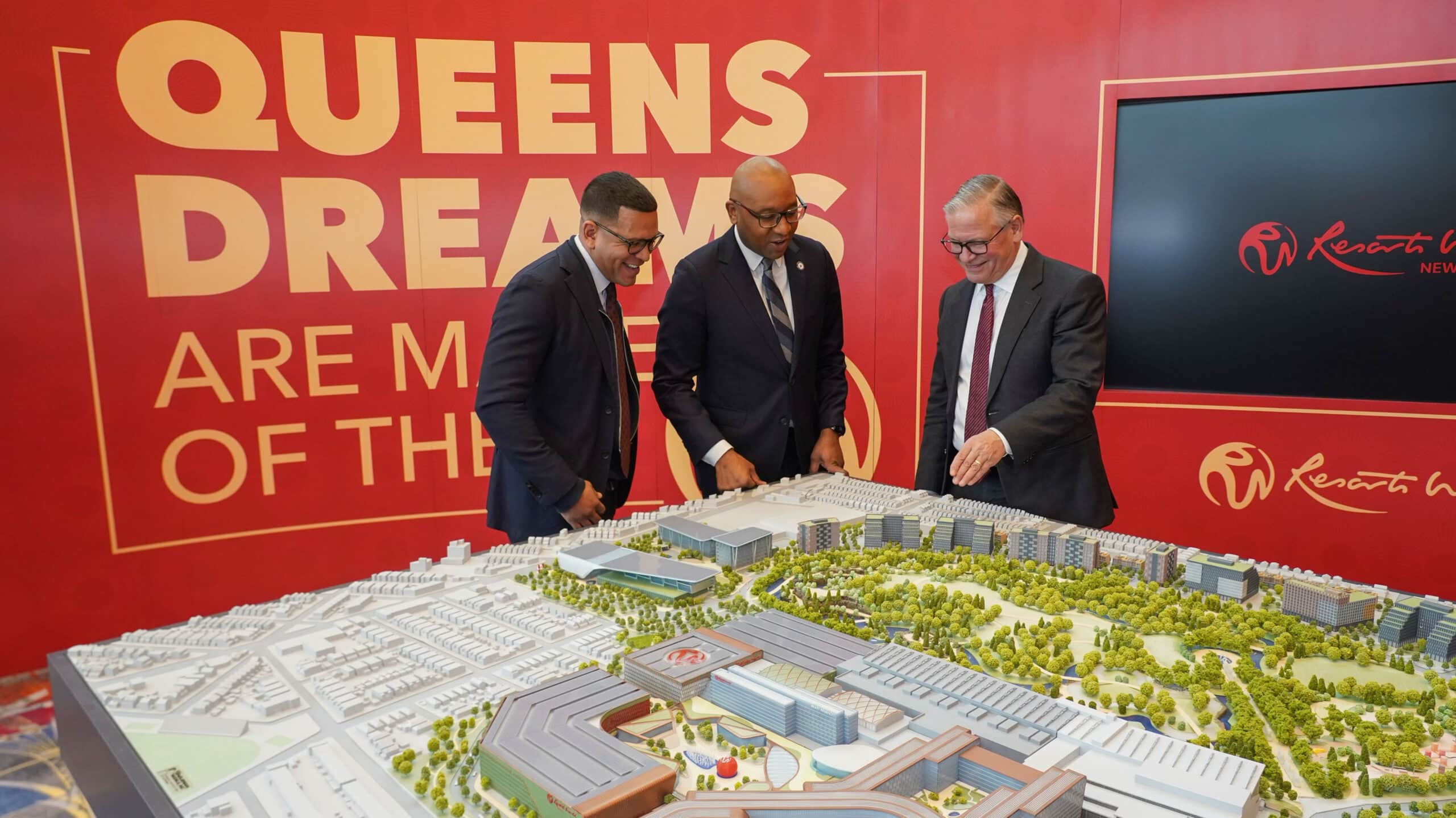 Three businessmen examining a detailed model of a neighborhood with the text "Queens Dreams are made of this" displayed above.