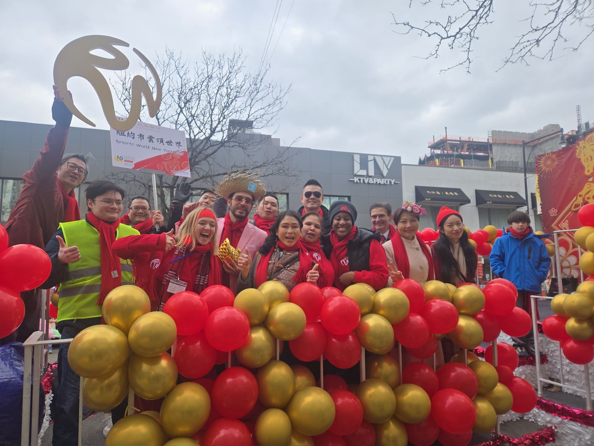 A group of people wearing red and yellow clothing and holding balloons, standing in front of a building with an "LVY Party" sign.