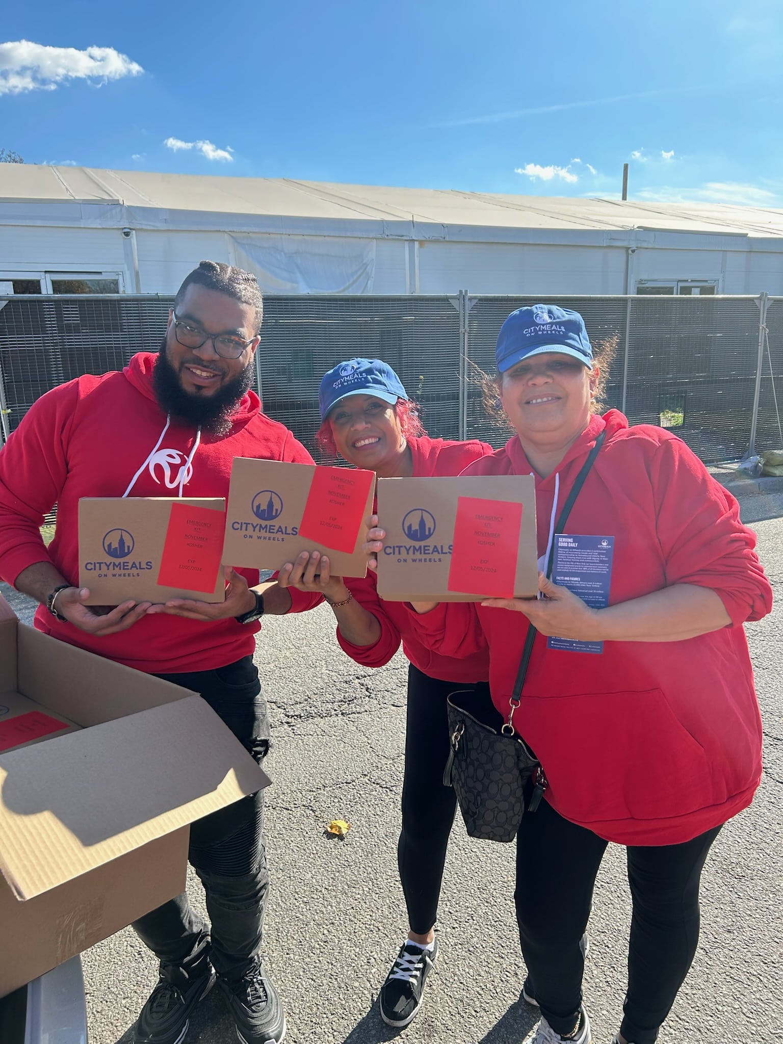 Three people wearing red shirts with "City Meals on Wheels" logos holding food delivery boxes and smiling.