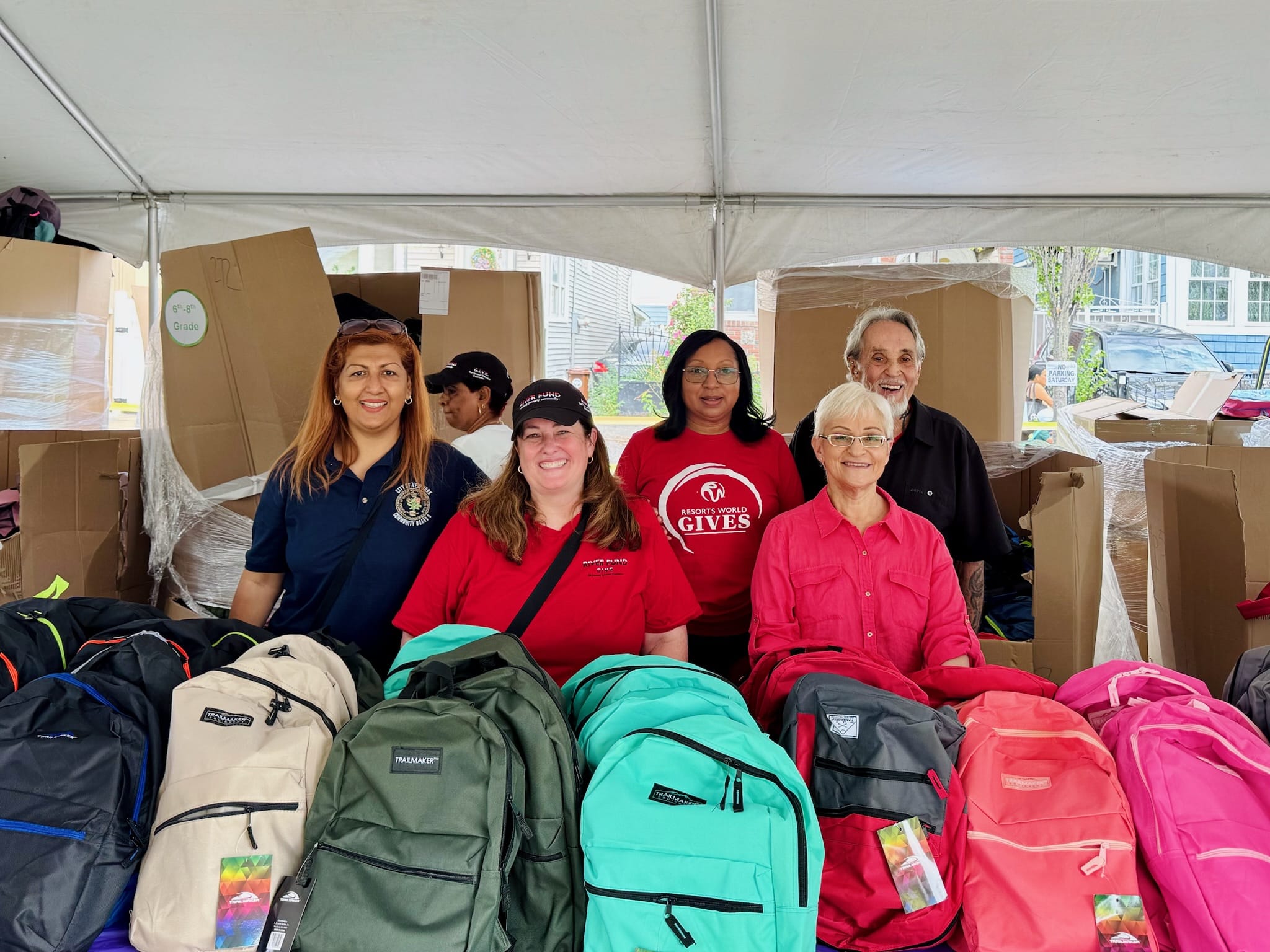A group of smiling volunteers in red shirts stand beside boxes and backpacks. The sign on the shirts reads "Students Give"