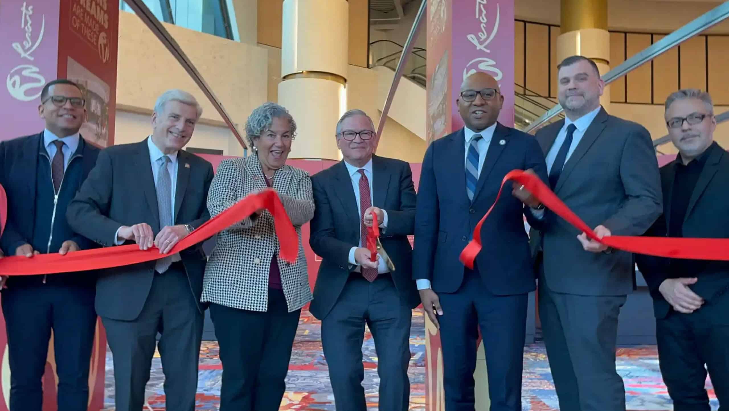 A group of 6 people in suits cutting a red ribbon to celebrate the opening of a new facility, with signs and banners in the background.