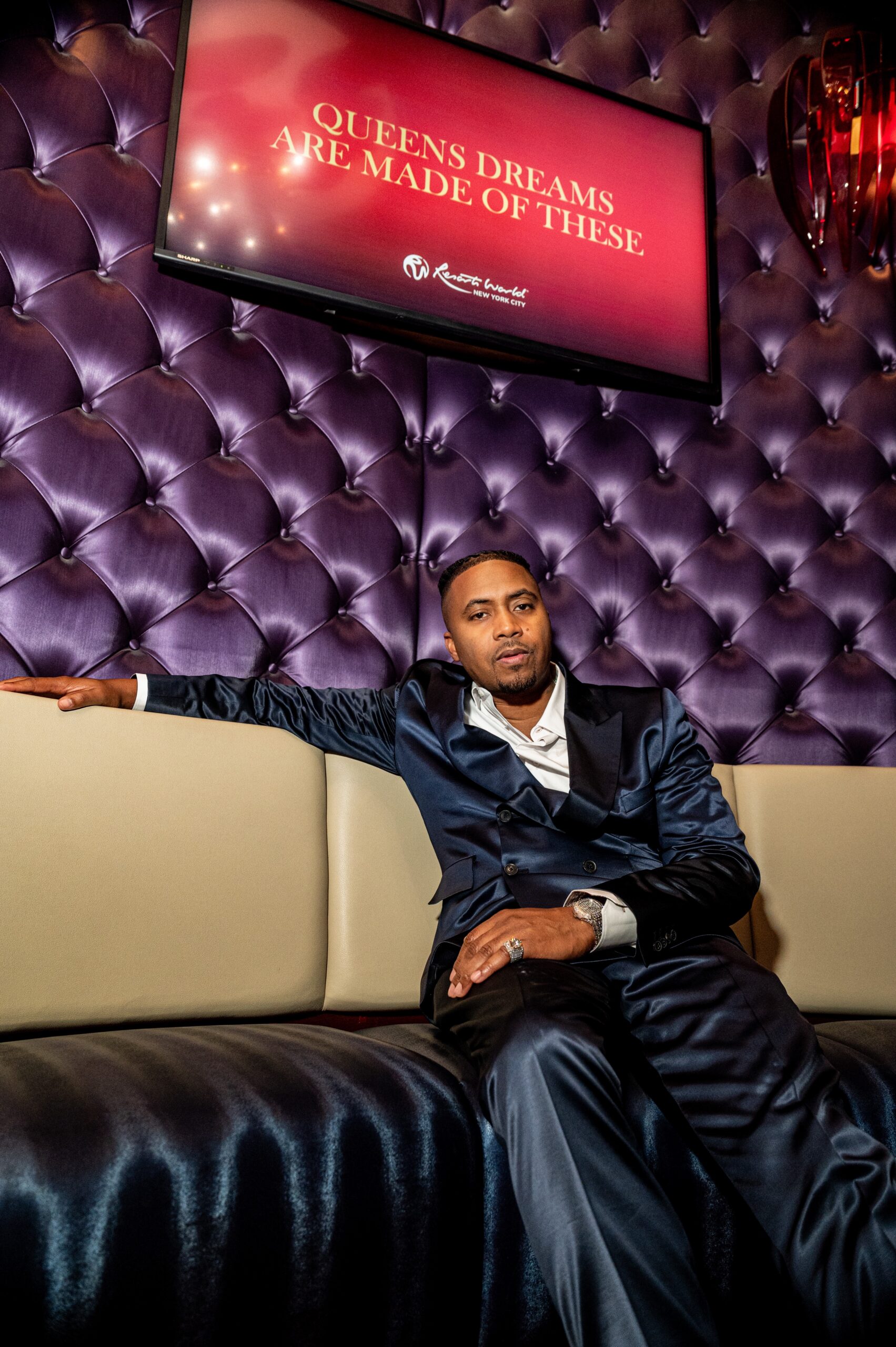 A man in a suit sits on a couch in front of a large sign that reads "QUEENS DREAMS ARE MADE OF THESE".