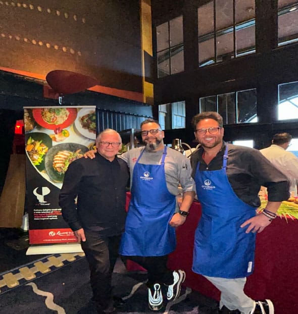 Three men in aprons standing together in a restaurant kitchen, surrounded by kitchen equipment and menus.