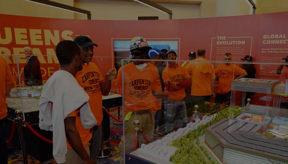 A group of people wearing orange "Carpenter" shirts in front of a large background of information and exhibits about the Queens development.