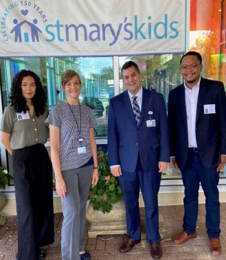 Four people standing in front of a "St. Mary's Kids" banner at what appears to be a conference or event.