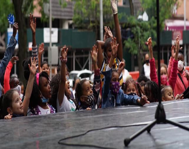A crowd of people, mostly children, standing with their hands raised in an energetic celebration or performance.