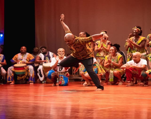 A lively performance featuring dancers in colorful traditional African attire performing on stage in front of a large audience.