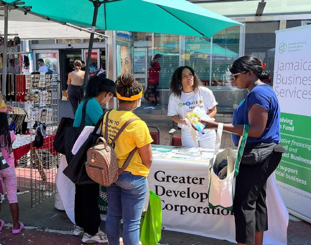 Outdoor public event booth with "Greater Development Corporation" banner and staff engaging with visitors.