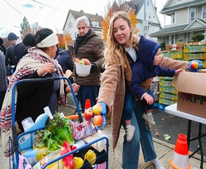 A group of people, including a woman with a baby stroller, shopping at an outdoor market or produce stand on a city street.