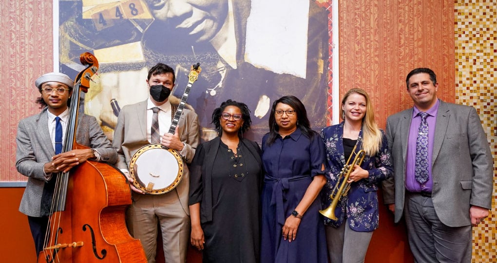 A diverse group of musicians posing together in front of an abstract mural backdrop, including a cellist, guitarist, two singers, a trumpet player, and a banjo player.