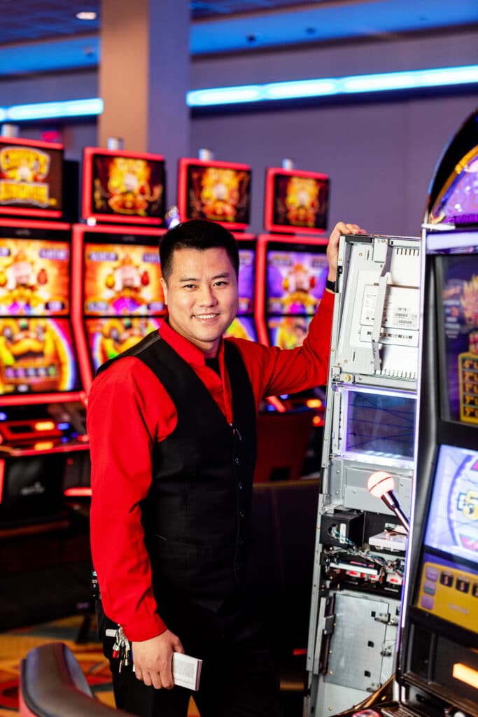 A smiling Asian man wearing a red and black uniform standing in front of video slot machines in a gaming arcade.