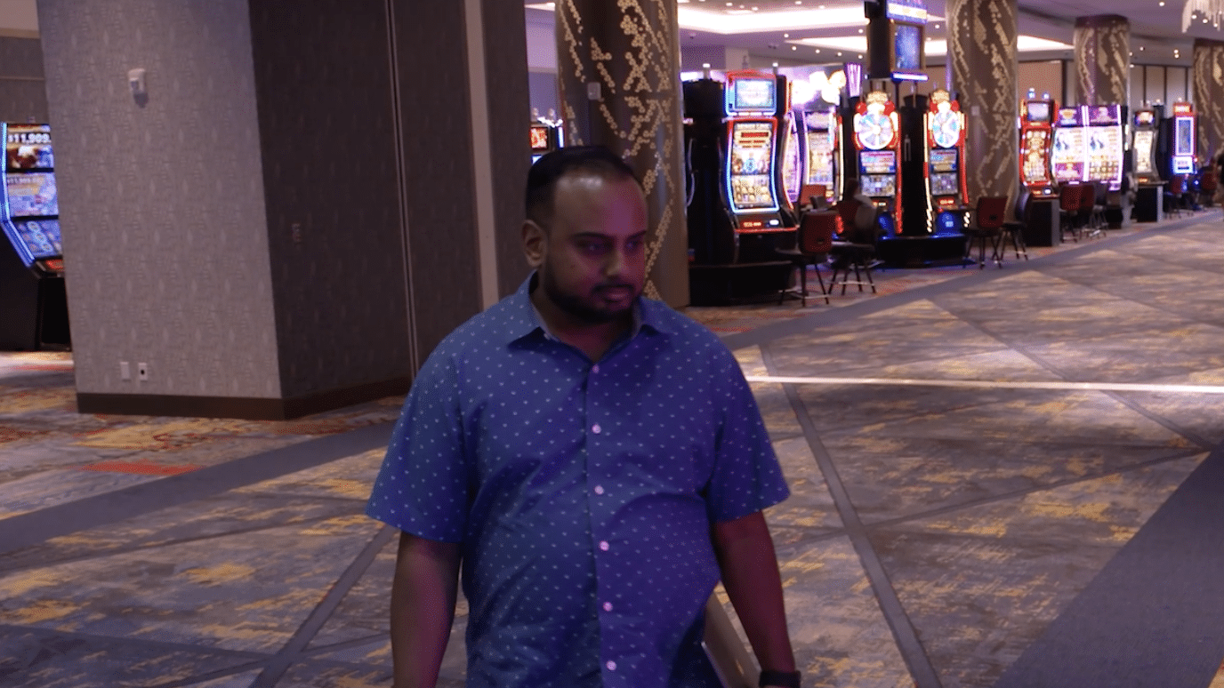 A man in a blue patterned shirt walks through a casino with slot machines visible in the background.