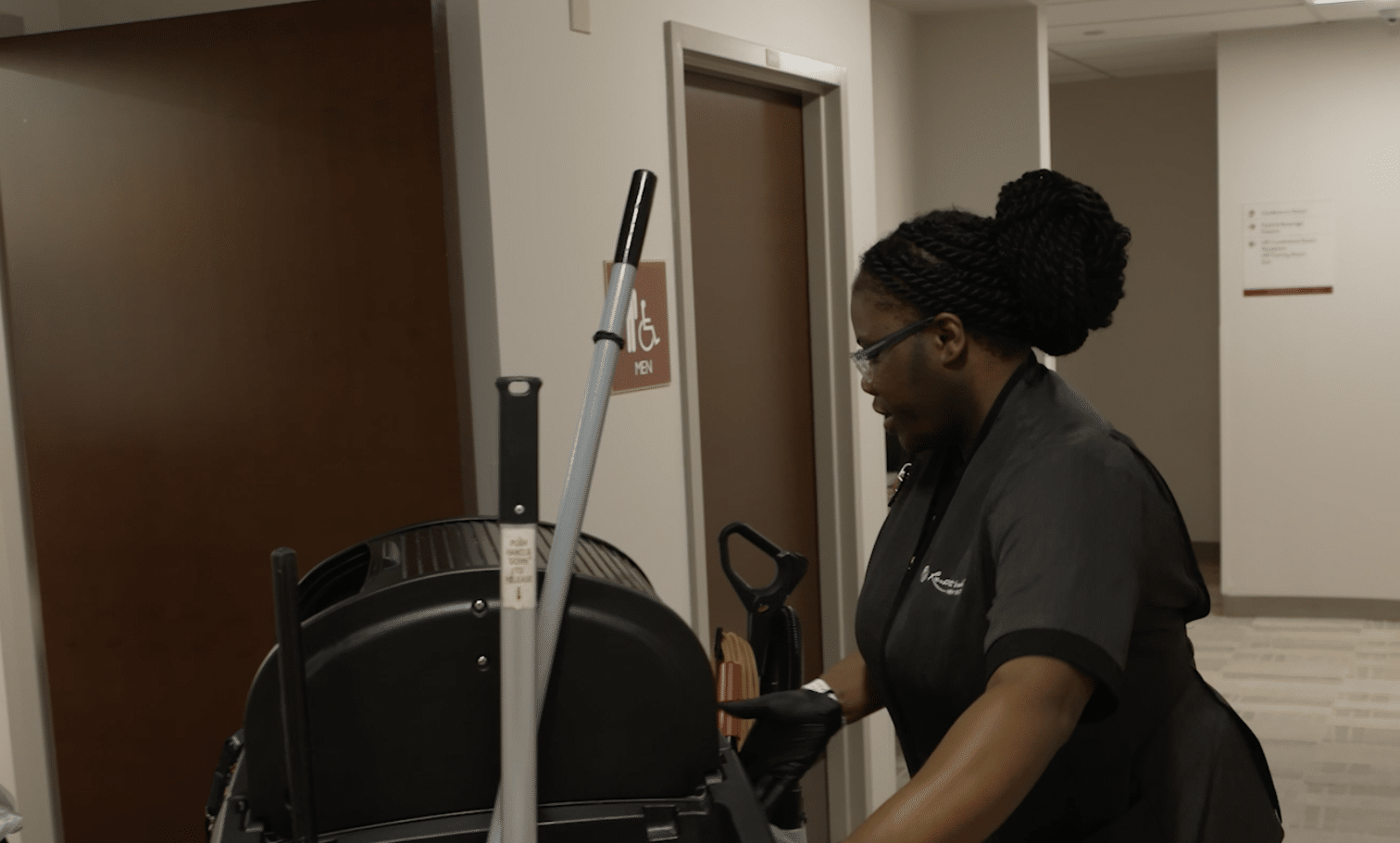 A person in a dark uniform pushing a large, black cleaning machine through a hallway.