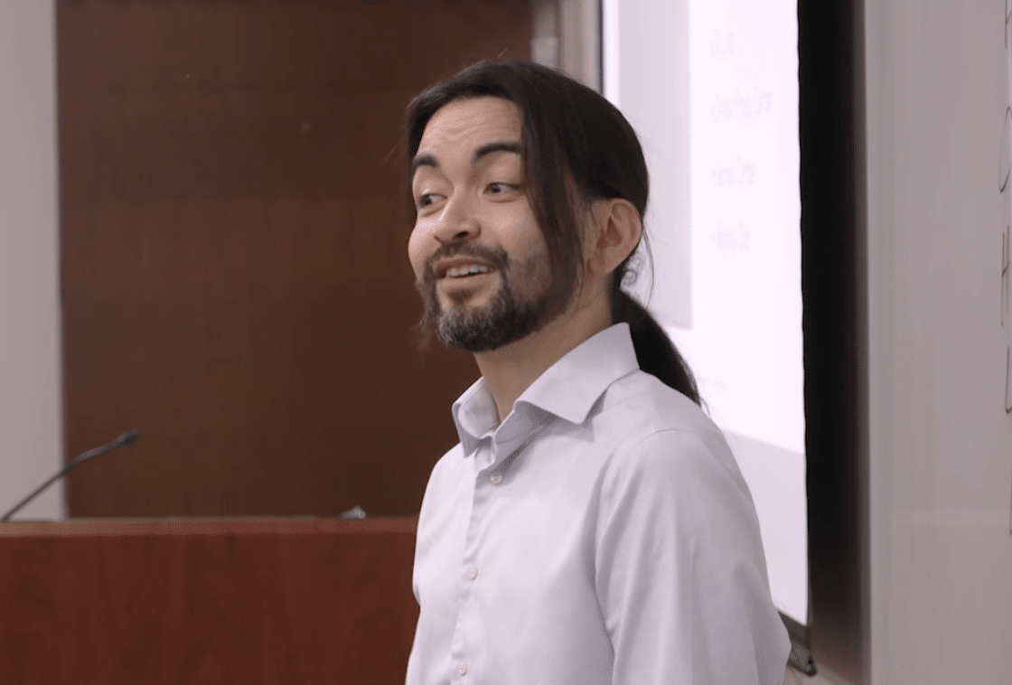 A man in a white shirt and beard standing in a room, smiling.