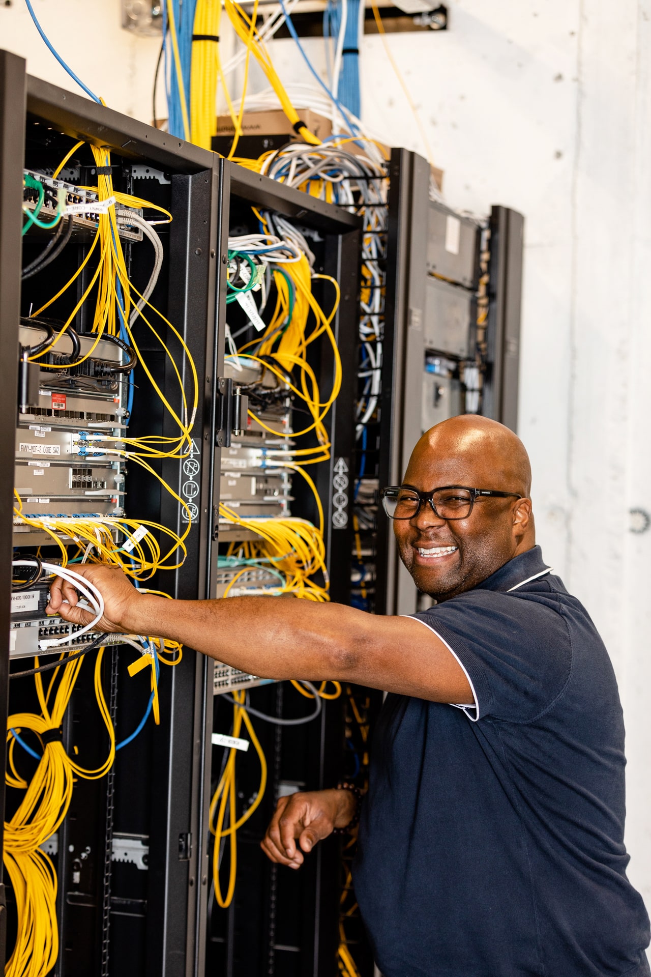 A smiling Black IT technician working on a server rack with many colored cables and wires.