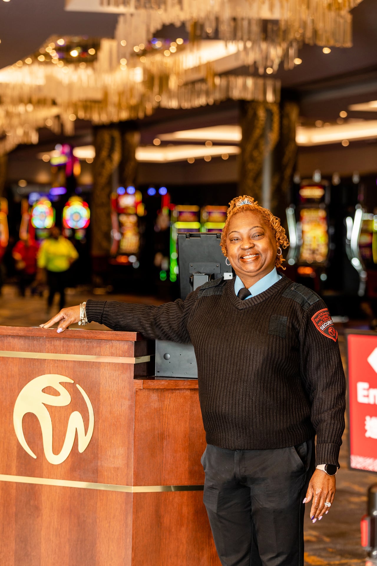 A smiling Black woman wearing a security uniform stands behind a wooden service counter in a brightly lit commercial space with arcade games and other entertainment equipment.