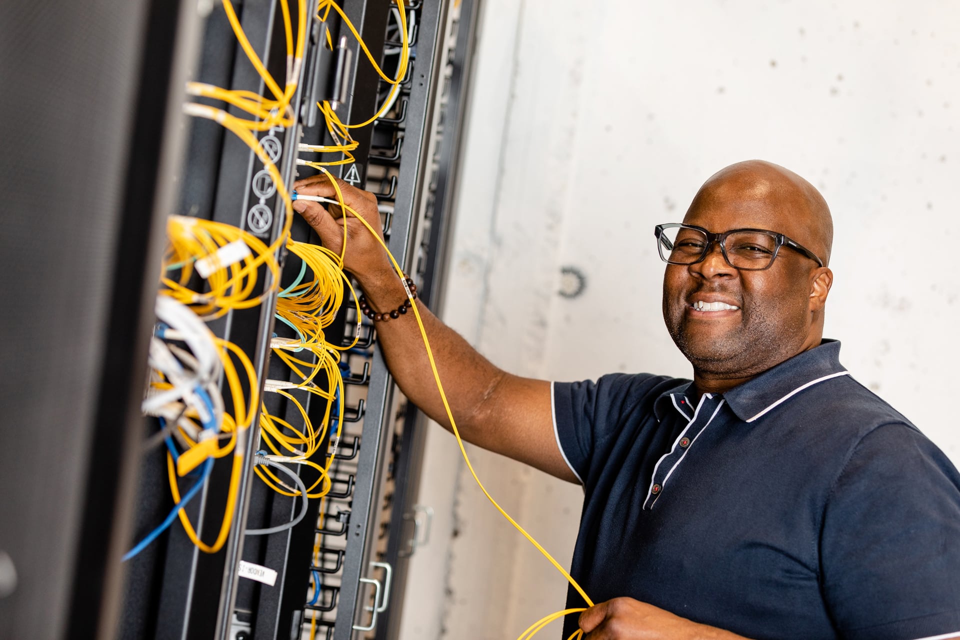 A smiling Black man wearing glasses and a navy blue shirt stands in front of a data center, working on a tangle of yellow cables.