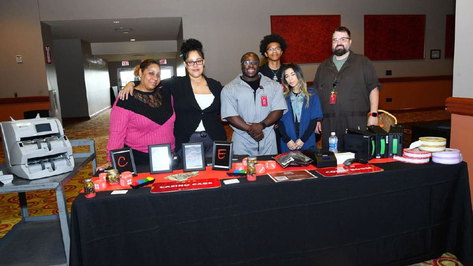 A group of six people, diverse in age and ethnicity, standing behind a table displaying casino-related items including cards, chips, and electronics. The table has "CASINO GAME" written on it.